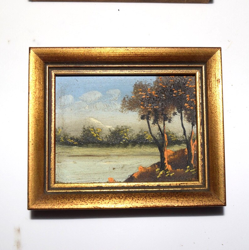 Op de afbeelding: Kleine ingelijste landschapsschilderij met een goudkleurige lijst. De schildering toont een serene sc&egrave;ne met een rivier, bomen en een bergketen onder een blauwe lucht met wolken. Het kunstwerk is waarschijnlijk een olieverfschilderij.