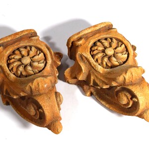 May include: Two antique, carved wooden architectural ornaments. Each piece features a central floral design and ornate scrollwork, finished in a warm, natural wood tone. These decorative elements are likely for interior design or restoration projects.