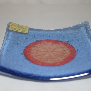 May include: A small, square, blue glass dish with a red circle in the center. The circle has a brown, floral design inside. The dish has a label that reads "Arte Viva - Via Ammendola 14 Cagliari".