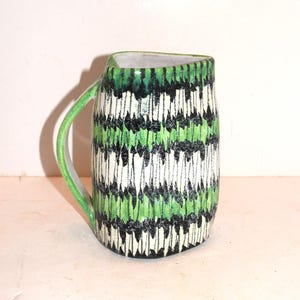 May include: A ceramic pitcher featuring a repeating vertical pattern of black and bright green brushstrokes over a cream-colored base. The vessel has a curved handle on the left side and a wide, slightly triangular opening at the top rim.