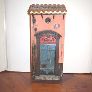 May include: A miniature wall-mounted facade featuring a salmon-colored exterior with distressed paint, a blue double door with a red arched transom, a brown tiled roof, and the number 83 displayed on the side. The structure includes a drainpipe.