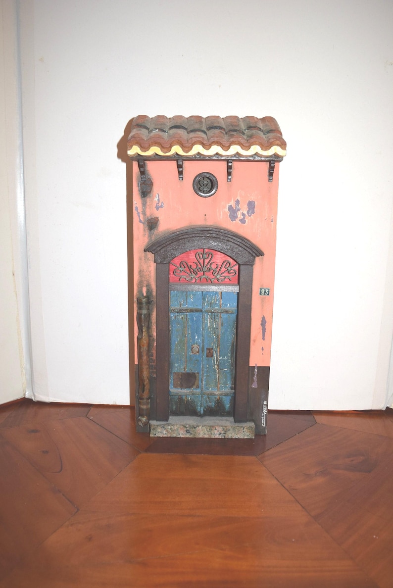May include: A miniature wall-mounted facade featuring a salmon-colored exterior with distressed paint, a blue double door with a red arched transom, a brown tiled roof, and the number 83 displayed on the side. The structure includes a drainpipe.
