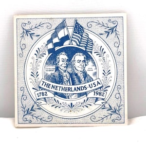 May include: Square ceramic tile with a blue and white design. The tile features portraits of two men, flags of the Netherlands and the USA, and the text "THE NETHERLANDS-USA 1782-1982". Decorative floral border.