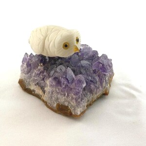 May include: A small, carved white owl figurine with yellow eyes resting on top of a jagged, deep purple amethyst crystal cluster. The owl has detailed feather carvings across its body and a smooth, rounded shape, contrasting with the sharp, angular points of the raw purple mineral base.