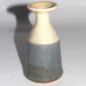 May include: A small ceramic vase with a flared rim and a tapered neck. The upper portion is a speckled off-white, while the lower body features a gradient of textured blue-gray tones. The base is cylindrical with a smooth, glazed finish.