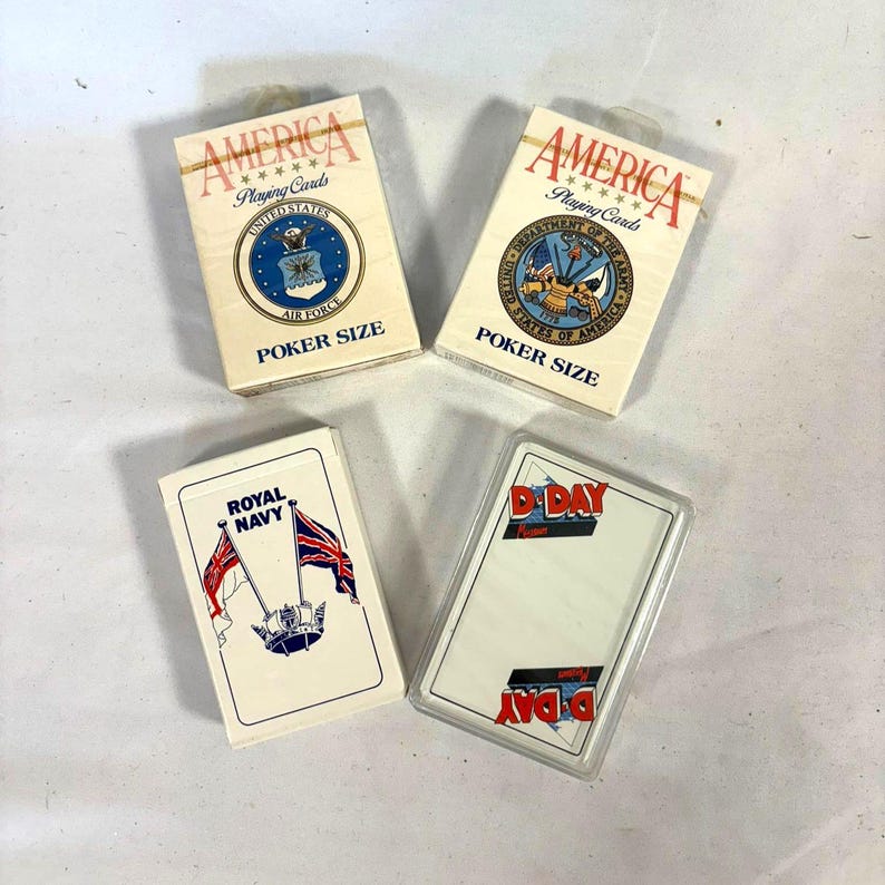 Puede incluir: Cuatro barajas de cartas de p&oacute;quer. Dos barajas est&aacute;n etiquetadas como "AMERICA" con emblemas militares y "POKER SIZE". Una baraja es "ROYAL NAVY" con banderas. La &uacute;ltima baraja est&aacute; etiquetada como "D-DAY".