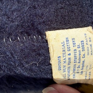 May include: Close-up of a navy blue plush fabric with white stitching and a cream-colored tag. The tag reads "ALL NEW MATERIAL SYNTHETIC FOAM & COTTON CALIFORNIA STUFFED TOYS" with an address and state abbreviations.