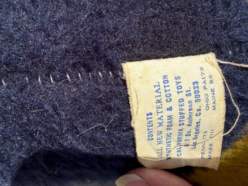 May include: Close-up of a navy blue plush fabric with white stitching and a cream-colored tag. The tag reads "ALL NEW MATERIAL SYNTHETIC FOAM & COTTON CALIFORNIA STUFFED TOYS" with an address and state abbreviations.