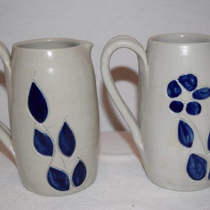 May include: Two off-white ceramic pitchers with curved handles. One pitcher features a blue floral design, while the other has a blue leaf pattern. Both pitchers have a smooth, glossy finish and a classic, functional design.