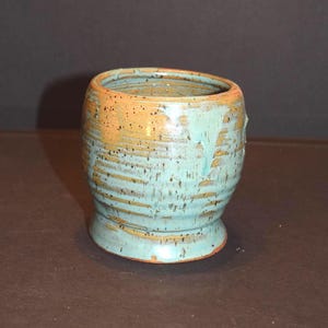 May include: A ceramic cup with a rounded body and a flared base, featuring a glossy glaze in shades of light blue and warm orange. The surface displays horizontal ridges and dark speckles throughout, with a drip effect near the rim.