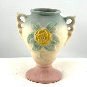 May include: A ceramic vase featuring a pastel color palette with a light pink base, a cream-colored body, and a light blue rim. The front displays a raised, embossed yellow flower with light green leaves. It has two decorative side handles.