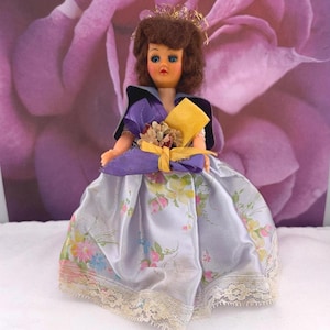 Könnte beinhalten: Eine Vintage-Puppe mit braunen Haaren, blauen Augen und einer goldenen Krone. Die Puppe trägt ein lila-schwarzes Outfit mit einem floral gemusterten Rock. Sie hält einen gelben Blumenstrauß, der mit einem lila Band gebunden ist. Der Rock hat eine Spitzenborte.