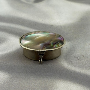 May include: An oval-shaped metal pill box featuring an iridescent abalone shell inlay on the lid. The container has a smooth, metallic finish and a small, hinged latch mechanism on the front for opening and closing the compact storage vessel.