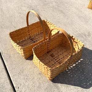 May include: Two woven rectangular baskets with arched handles. The baskets are made of light brown woven material and sit on a concrete surface. The baskets are empty and appear to be handmade.