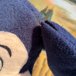 May include: A close-up of a plush toy featuring a cartoon character's face and ear. The toy has a navy blue body and ear, with a beige face and black details. The toy appears to be well-loved.