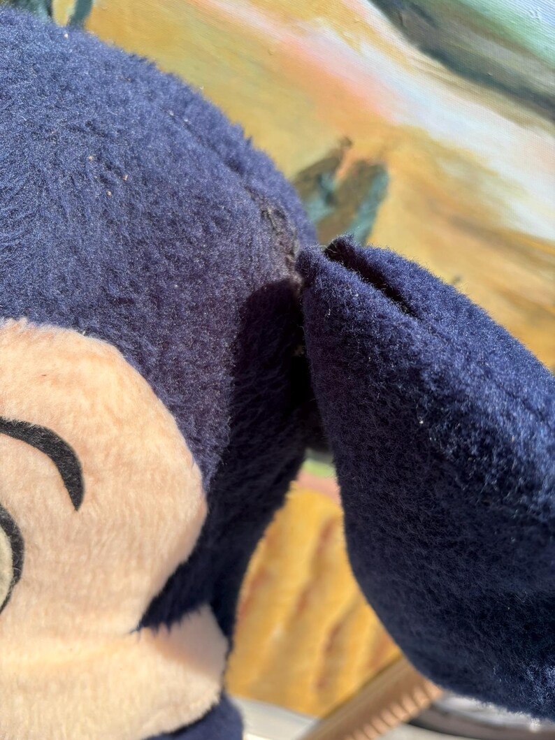 May include: A close-up of a plush toy featuring a cartoon character's face and ear. The toy has a navy blue body and ear, with a beige face and black details. The toy appears to be well-loved.