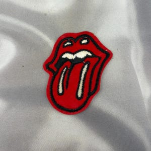 May include: An embroidered patch featuring a stylized red mouth with thick black outlines and white teeth. The design depicts a pair of lips with a tongue protruding downwards, rendered in vibrant red thread with black and white detailing.