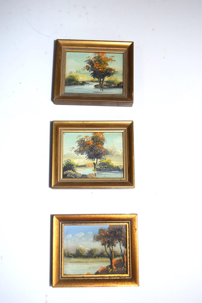 Op de afbeelding: Drie kleine ingelijste landschapsschilderijen. Elk toont een boom met oranje en bruine bladeren in de buurt van water. De goudkleurige frames en schilderijen worden tentoongesteld tegen een witte achtergrond.