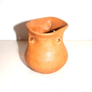 May include: A small, hand-formed terracotta pot with a rounded body and a flared, uneven rim. The vessel features two small, circular holes positioned symmetrically on the upper sides, just below the opening, with a smooth, matte finish.