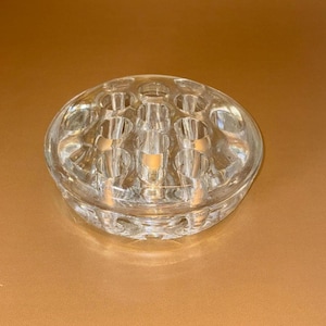 May include: A clear, circular glass flower frog featuring multiple cylindrical openings arranged in a grid pattern. This heavy, transparent glass piece is designed with a smooth, rounded exterior edge and a flat base for stability in floral arrangements.