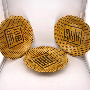 May include: Three decorative yellow plates with a scalloped edge. Each plate features a black geometric design in the center. The plates are arranged on a white surface.