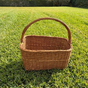 May include: A rectangular brown wicker basket with a woven handle. The basket is sitting on a bed of green grass, with a hedge in the background. The basket is empty and appears to be new.