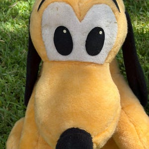 May include: A plush toy of Pluto, a yellow dog with black ears and a black nose. The toy has large black eyes with white eye surrounds. The plush is sitting on green grass.