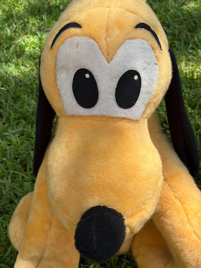 May include: A plush toy of Pluto, a yellow dog with black ears and a black nose. The toy has large black eyes with white eye surrounds. The plush is sitting on green grass.