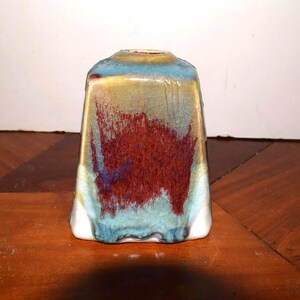 May include: A square-shaped ceramic vase with a tapered top and a circular opening. The glaze features a crackle finish with a blend of deep red, turquoise, and golden-yellow tones, creating a multi-colored, textured appearance on the surface.