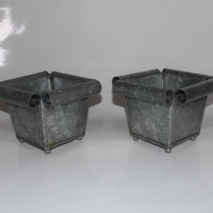 May include: Two gray metal planters with rolled edges and decorative scrollwork. The planters have a square base and are sitting on a white surface.