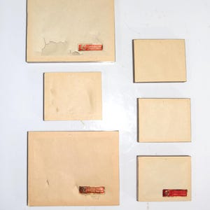 Op de afbeelding: Een verzameling vintage, rechthoekige kaarten in verschillende maten. De kaarten zijn lichtbeige van kleur, sommige vertonen tekenen van slijtage, waaronder afbladderende randen. Elke kaart heeft een klein, rood rechthoekig label met tekst.