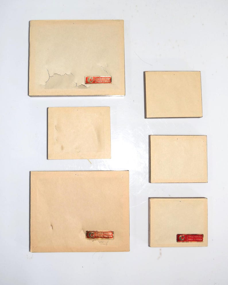 Op de afbeelding: Een verzameling vintage, rechthoekige kaarten in verschillende maten. De kaarten zijn lichtbeige van kleur, sommige vertonen tekenen van slijtage, waaronder afbladderende randen. Elke kaart heeft een klein, rood rechthoekig label met tekst.