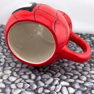 May include: A red ceramic mug featuring a Spider-Man design. The mug is red with black web lines and a white and black mask detail. It has a red handle and a beige interior.