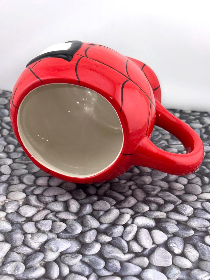May include: A red ceramic mug featuring a Spider-Man design. The mug is red with black web lines and a white and black mask detail. It has a red handle and a beige interior.