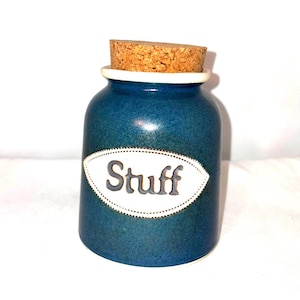 May include: A small, blue ceramic jar with a cork stopper. The jar has a white oval label with the word "Stuff" in gray lettering. The jar is designed to hold small items.