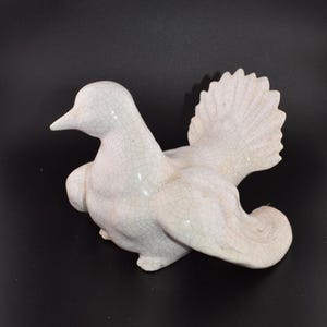 May include: A white ceramic dove figurine with outstretched wings. The bird has a detailed, cracked glaze finish. The dove is positioned on a black surface, showcasing its form and texture. A decorative piece for home decor.