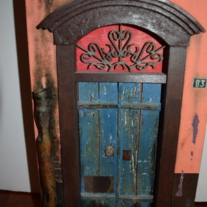 May include: A weathered wooden door with a blue paint finish and a metal grate above it. The door is framed in dark wood and set into a wall with a pink and orange paint finish. The number 83 is visible on the wall.