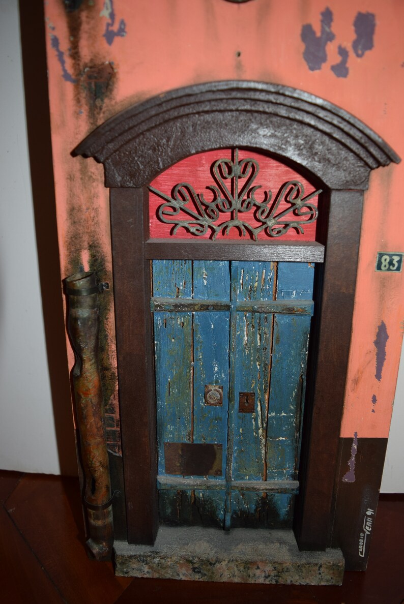 May include: A weathered wooden door with a blue paint finish and a metal grate above it. The door is framed in dark wood and set into a wall with a pink and orange paint finish. The number 83 is visible on the wall.
