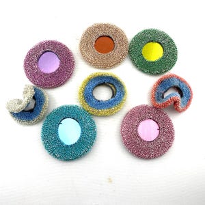 May include: A collection of colorful, circular, and textured decorative items. The items feature a variety of colors, including pink, green, blue, and gold. Some have a central, contrasting color, while others are folded or curved.