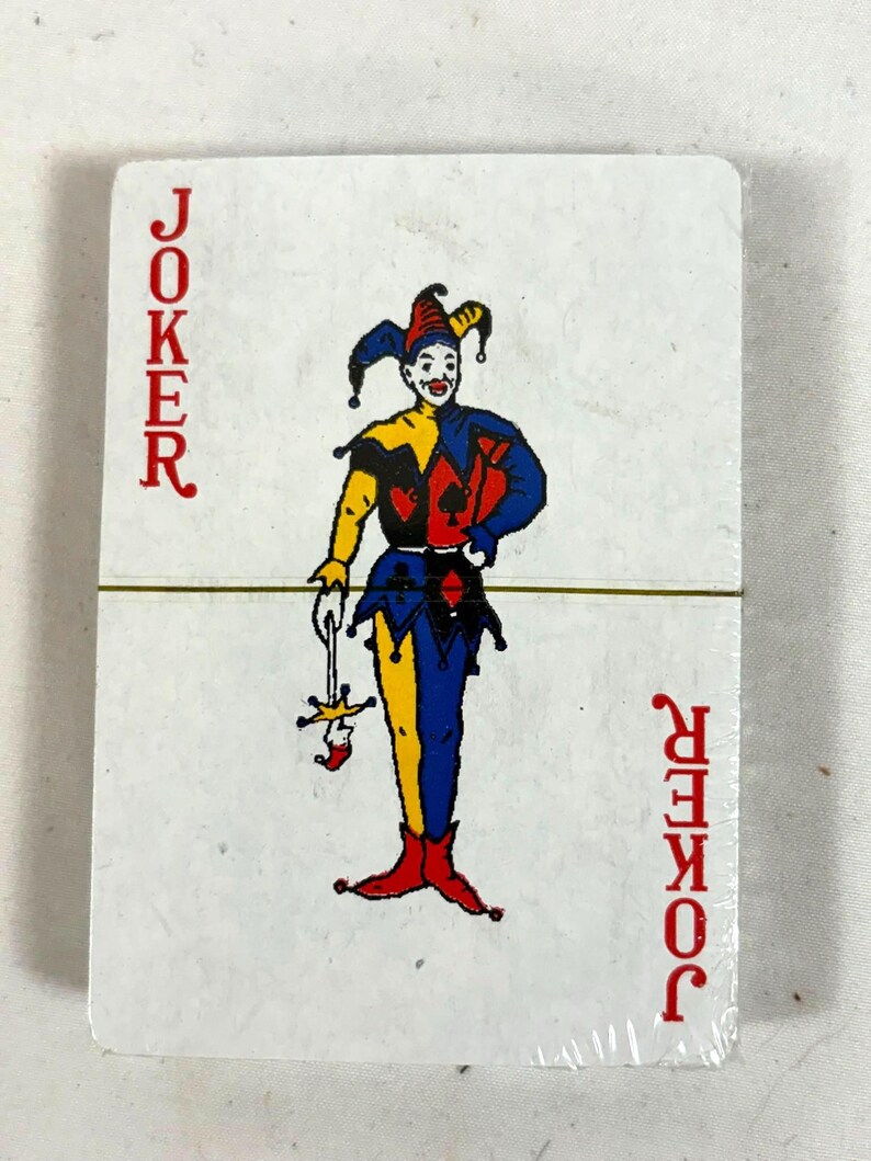 Puede incluir: Un mazo de cartas con una carta de Joker. La carta muestra un buf&oacute;n colorido con un atuendo rojo, amarillo, azul y negro. La palabra "JOKER" est&aacute; impresa verticalmente en rojo en ambos lados de la carta.