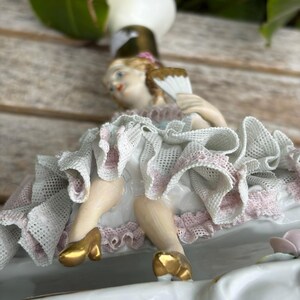 May include: A porcelain figurine of a woman in a white dress with pink trim, holding a fan. The figurine has gold shoes and is part of a decorative piece. The overall design is ornate and detailed, with a vintage aesthetic.