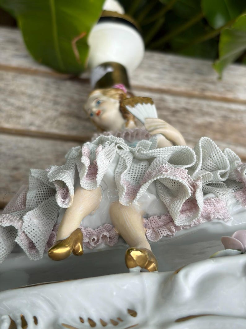 May include: A porcelain figurine of a woman in a white dress with pink trim, holding a fan. The figurine has gold shoes and is part of a decorative piece. The overall design is ornate and detailed, with a vintage aesthetic.