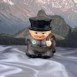 May include: A ceramic figurine of a person wearing a black top hat and a brown coat with an orange collar. The figure has a light green clover pinned to the chest and holds a brown circular object in one hand, featuring painted facial features.