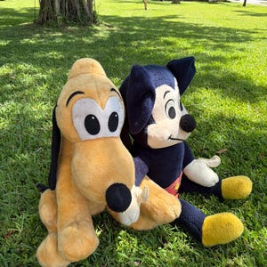 May include: Two plush Disney character toys, Pluto and Mickey Mouse, sit on green grass. Pluto is yellow with black ears and a black nose. Mickey Mouse is wearing a red and yellow outfit with black ears and a black nose.
