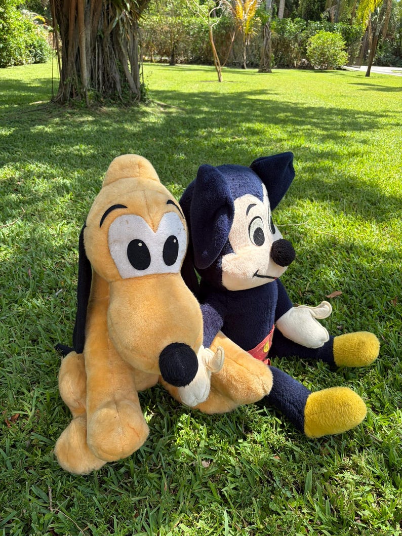 May include: Two plush Disney character toys, Pluto and Mickey Mouse, sit on green grass. Pluto is yellow with black ears and a black nose. Mickey Mouse is wearing a red and yellow outfit with black ears and a black nose.