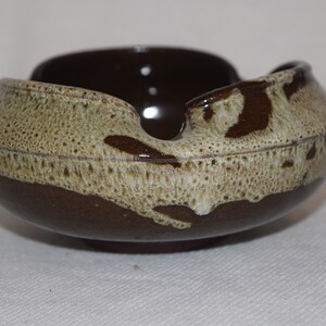 May include: A brown and white ceramic ashtray with a unique, wavy design. The ashtray has a speckled glaze and a wide, shallow bowl.