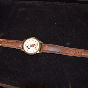 May include: A brown leather wristwatch with a white face featuring Mickey Mouse. The watch has black numbers and hands.