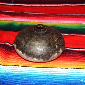 May include: A dark brown, round ceramic vessel with a small opening at the top. The vessel has a textured surface with subtle etched lines and a lighter-colored base. The background features a colorful striped textile.