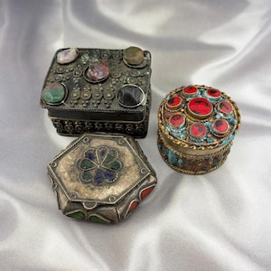 May include: Three ornate, antique metal boxes with gemstone embellishments. One is rectangular with a dark finish and various stones. Another is round with red stones and turquoise accents. The third is a silver hexagonal box with a floral design.