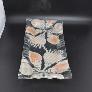 May include: Rectangular glass dish with a black background and a design of seashells in shades of white, beige, and orange. The edges of the dish have a wavy design. A decorative piece for the home.